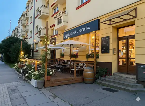 A wide-angle, photorealistic image of a quirky café on a cobblestone street in Žižkov, Prague. The café has a charming, bohemian vibe with mismatched wooden tables and chairs on an outdoor patio, adorned with string lights and abundant potted plants. A vintage bicycle rests against the wall, which features colorful, artistic graffiti. The warm, soft lighting suggests a welcoming atmosphere at dusk.